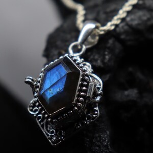 May include: A close-up of a silver pendant with a faceted, blue gemstone. The gemstone is set in an ornate, filigree-style setting. The pendant hangs from a silver chain, and the background is dark.