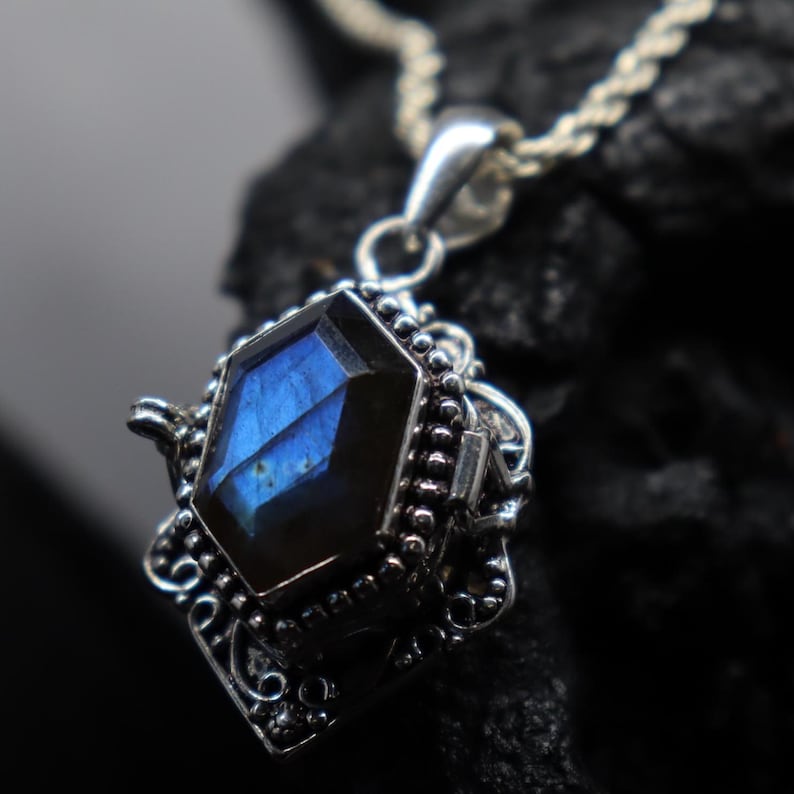 May include: A close-up of a silver pendant with a faceted, blue gemstone. The gemstone is set in an ornate, filigree-style setting. The pendant hangs from a silver chain, and the background is dark.