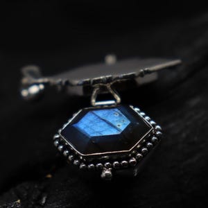 May include: A silver pendant featuring a faceted, hexagonal gemstone with blue and iridescent hues. The gemstone is set in a detailed, beaded silver frame, suspended from a stylized silver element. The pendant is displayed against a dark background.