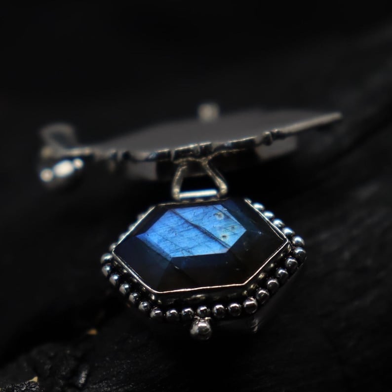May include: A silver pendant featuring a faceted, hexagonal gemstone with blue and iridescent hues. The gemstone is set in a detailed, beaded silver frame, suspended from a stylized silver element. The pendant is displayed against a dark background.