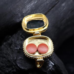 May include: A gold-colored, open locket pendant with a square shape and a hinged opening. Inside the locket are two small, round, reddish-brown objects. The locket is on a dark, textured surface.