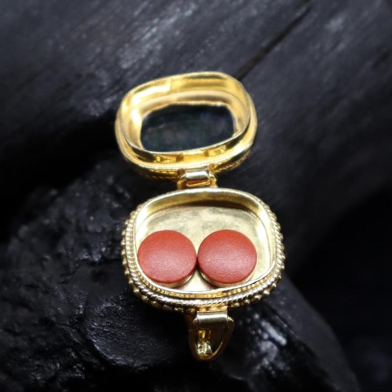 May include: A gold-colored, open locket pendant with a square shape and a hinged opening. Inside the locket are two small, round, reddish-brown objects. The locket is on a dark, textured surface.