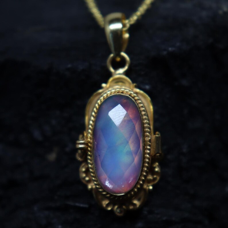 May include: A gold-colored pendant necklace featuring an oval-shaped, faceted gemstone with a blend of blue, purple, and pink hues. The pendant is intricately designed with ornate details and hangs from a delicate gold chain.