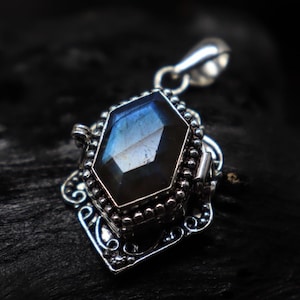 May include: A silver pendant with a large, faceted, blue-gray gemstone. The gemstone is set in a decorative, ornate silver frame with a bail for a chain. The pendant has a vintage or antique aesthetic.