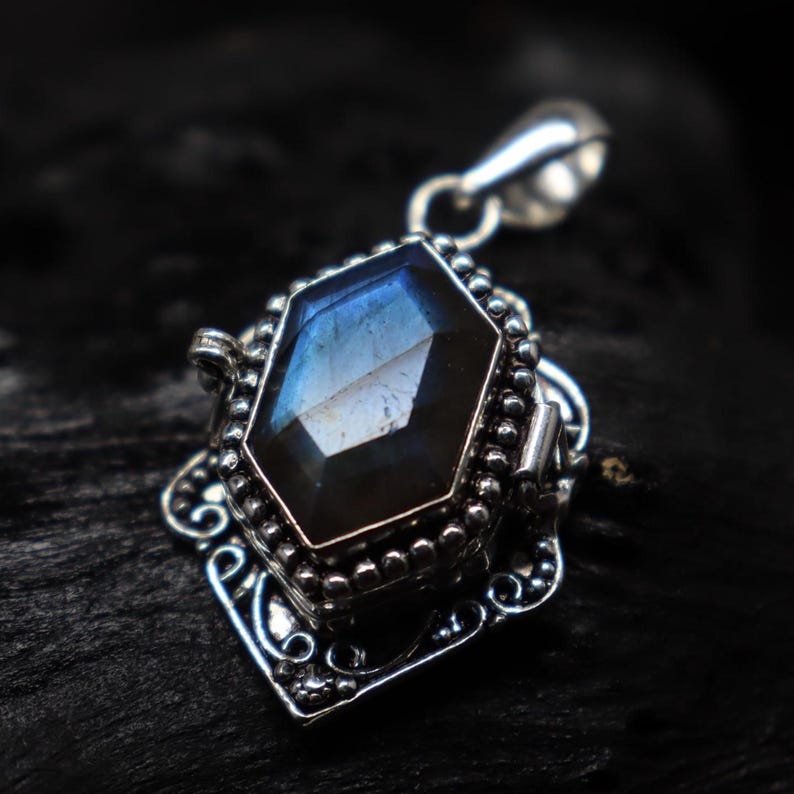 May include: A silver pendant with a large, faceted, blue-gray gemstone. The gemstone is set in a decorative, ornate silver frame with a bail for a chain. The pendant has a vintage or antique aesthetic.