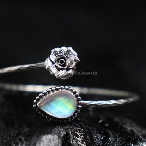 May include: An open cuff bracelet made of oxidized silver featuring a sculpted rose at one end and a pear-shaped, faceted iridescent gemstone set in a beaded frame at the other. The band has a textured, twisted metal finish throughout its length.