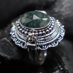 May include: A silver ring featuring a large, faceted, oval-shaped green gemstone. The ring has a detailed, ornate setting with a beaded border and decorative filigree around the gemstone. The ring is set against a dark background.