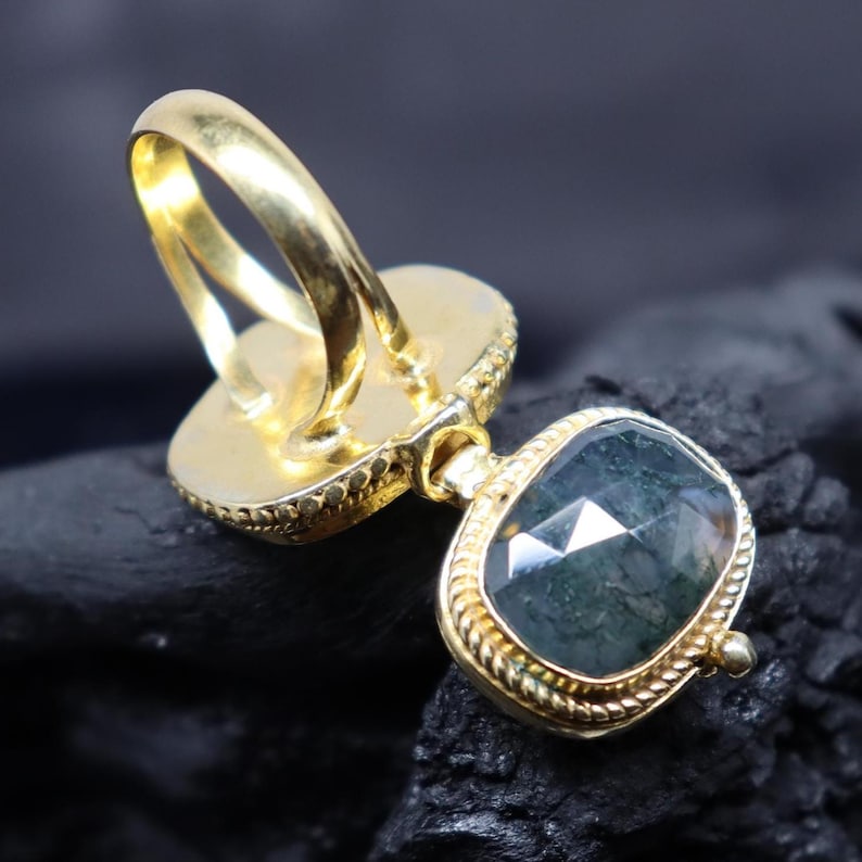 May include: A gold-colored ring with a large, faceted, dark green gemstone. The ring has a detailed, textured band and setting. The gemstone is oval-shaped and set in a decorative bezel. The ring is displayed against a dark background.
