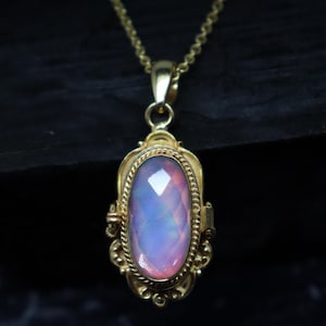 May include: A gold-colored pendant necklace featuring an oval, faceted gemstone with pink and blue hues. The pendant is set in a decorative frame with intricate detailing and hangs from a delicate chain. The necklace is displayed against a dark background.