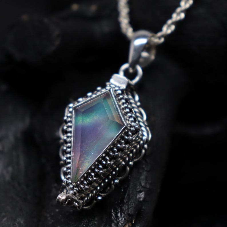 Pu&ograve; includere: Una collana con pendente in argento con una gemma geometrica e iridescente. Il pendente ha una forma a diamante con un bordo argentato dettagliato con perline. La gemma mostra sfumature verdi, viola e blu ed &egrave; sospesa a una catena d'argento.