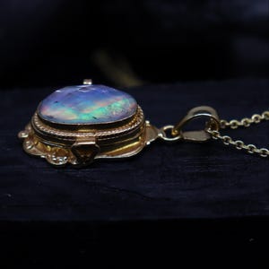 May include: A gold-colored pendant necklace featuring a round, iridescent opal gemstone. The gemstone is set in a detailed, ornate gold setting with a hinged lid. The necklace has a delicate gold chain.