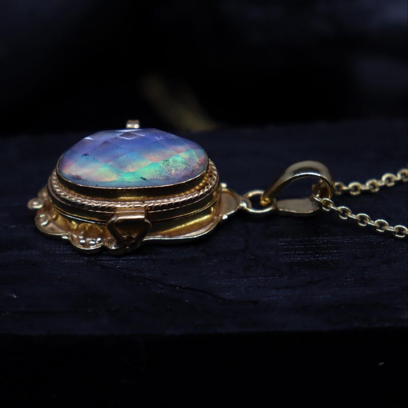 May include: A gold-colored pendant necklace featuring a round, iridescent opal gemstone. The gemstone is set in a detailed, ornate gold setting with a hinged lid. The necklace has a delicate gold chain.