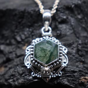 May include: A silver pendant necklace featuring a hexagonal green gemstone with intricate internal patterns. The gemstone is set in a detailed silver frame with decorative elements. The chain is a silver curb link.