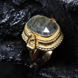 May include: A gold-colored ring with a large, rectangular, moss agate stone. The stone is a translucent green with dark inclusions. The ring has a detailed, ornate setting with a rope-like design around the stone.