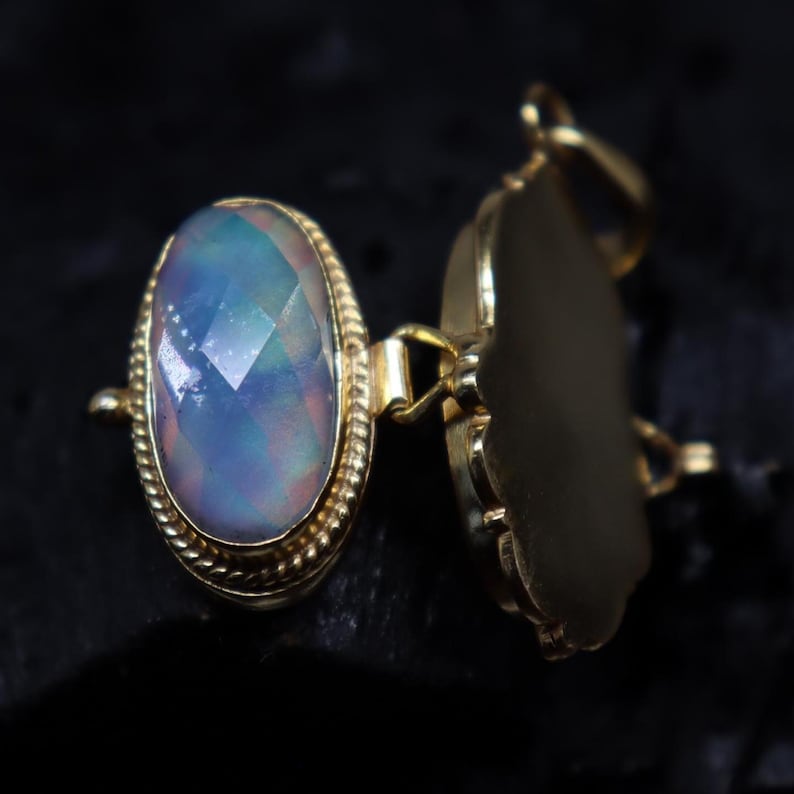 May include: A gold-colored locket with an oval, iridescent gemstone. The gemstone has a faceted cut, displaying shades of blue, pink, and green. The locket is open, revealing a blank interior. The setting has a rope-like border.