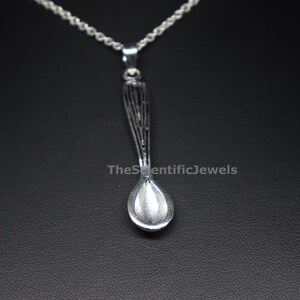 Op de afbeelding: Een zilverkleurige lepelhanger aan een bijpassende ketting. De lepel heeft een gestructureerd handvat met verticale lijnen en een gladde, ronde kom. De tekst TheScientificJewels is zichtbaar in het midden van de afbeelding over de lepel heen.