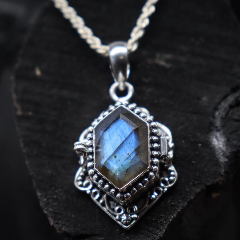 May include: A silver pendant necklace featuring a faceted, blue labradorite gemstone. The gemstone is set in an ornate, filigree-style frame. The necklace has a delicate, twisted chain.