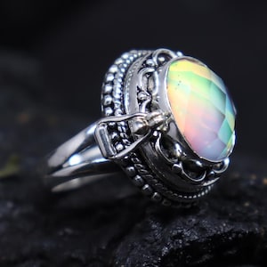 Op de afbeelding: Een zilveren ring met een ovale, iriserende edelsteen. De edelsteen toont een spectrum aan kleuren, waaronder geel, groen en blauw. De ring heeft ingewikkelde details rond de edelsteen en op de band. De ring staat tegen een donkere achtergrond.