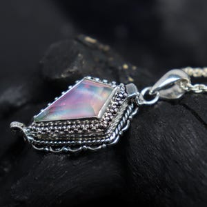 Pu&ograve; includere: Una collana con pendente in argento con una gemma triangolare iridescente. La gemma mostra sfumature di rosa, viola e verde. Il pendente ha un'incastonatura in argento dettagliata e ornata con un bordo decorativo.