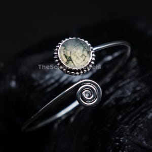 May include: A silver bypass ring featuring a round, bezel-set moss agate stone with dark green dendritic inclusions and a spiral terminal. The metal band has a beaded texture around the stone setting and the text TheSciencJewels is visible.
