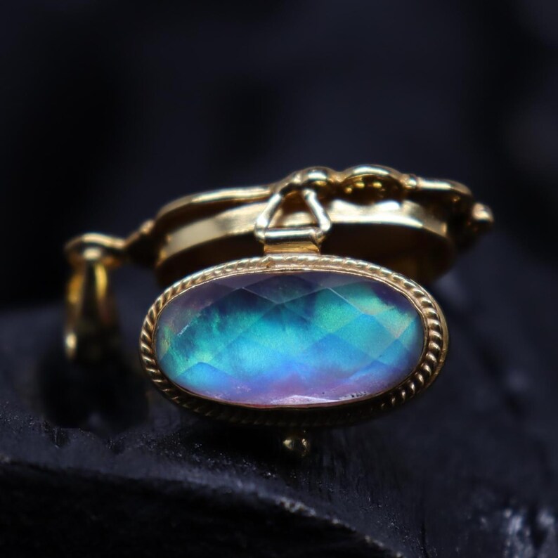May include: A gold-colored pendant with an oval, iridescent gemstone. The gemstone displays a range of colors, including blue, green, and purple. The pendant has a decorative, rope-like border around the gemstone and a detailed bail.