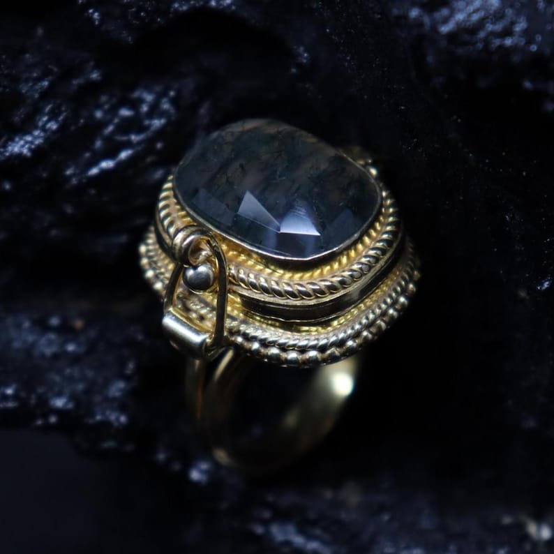 May include: A gold-toned ring with a large, faceted, dark gemstone. The ring features intricate detailing around the stone and along the band, with a textured, rope-like design. The gemstone has a rectangular shape with cut facets.