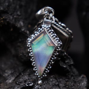 Pu&ograve; includere: Ciondolo in argento con una pietra preziosa iridescente a forma di diamante. La pietra preziosa mostra sfumature di blu, verde e rosa. Il ciondolo &egrave; incastonato in una cornice d'argento perlata e pende da un'ansa d'argento. Lo sfondo &egrave; scuro.