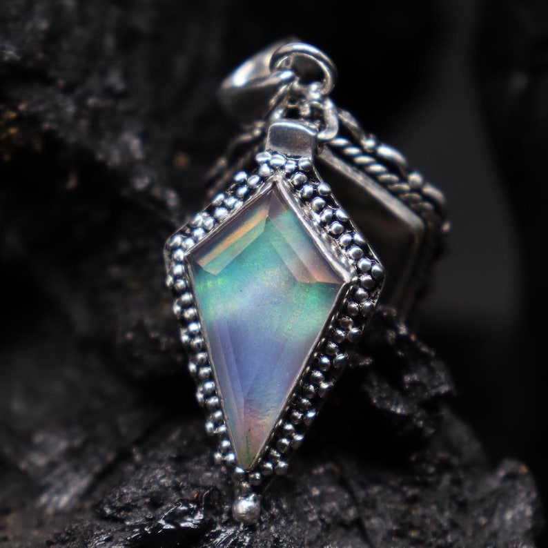 Pu&ograve; includere: Ciondolo in argento con una pietra preziosa iridescente a forma di diamante. La pietra preziosa mostra sfumature di blu, verde e rosa. Il ciondolo &egrave; incastonato in una cornice d'argento perlata e pende da un'ansa d'argento. Lo sfondo &egrave; scuro.