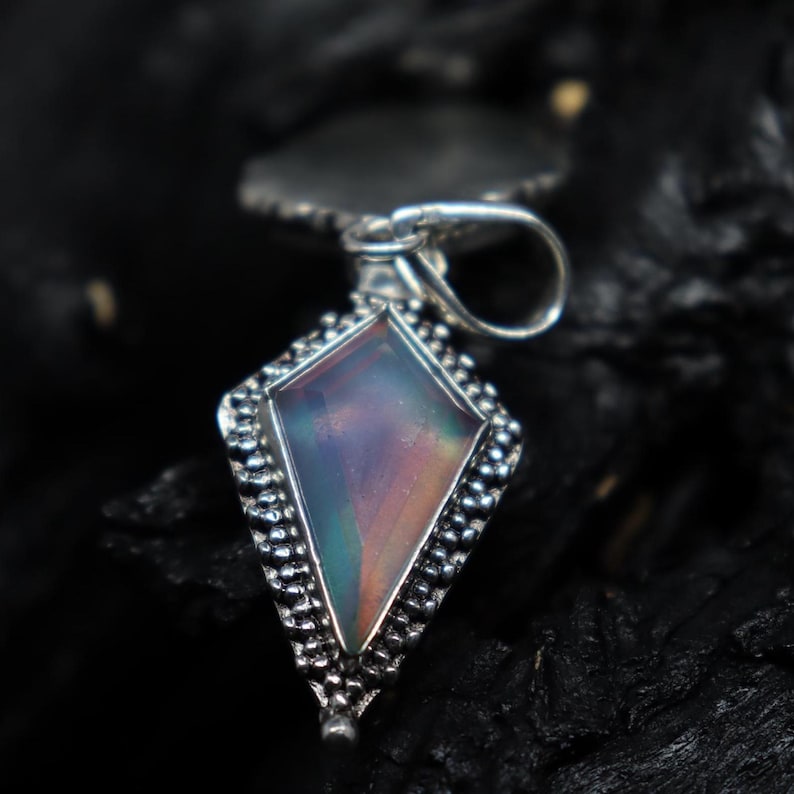 Pu&ograve; includere: Un ciondolo d'argento con una gemma iridescente a forma di diamante. La gemma mostra colori viola, blu e verde. Il ciondolo ha un bordo dettagliato con perline e un anello per una catena.