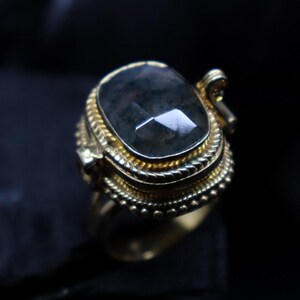 May include: A gold-toned ring with a large, faceted, dark green gemstone. The ring has intricate detailing around the stone and along the band, with a decorative element on the side. The ring is set against a dark background.