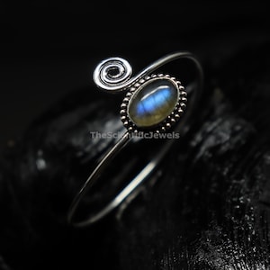 May include: A silver ring featuring an oval-shaped labradorite gemstone with a blue iridescent flash. The band has a spiral design on one end and a beaded setting around the stone. The text TheScientificJewels is visible on the ring band.