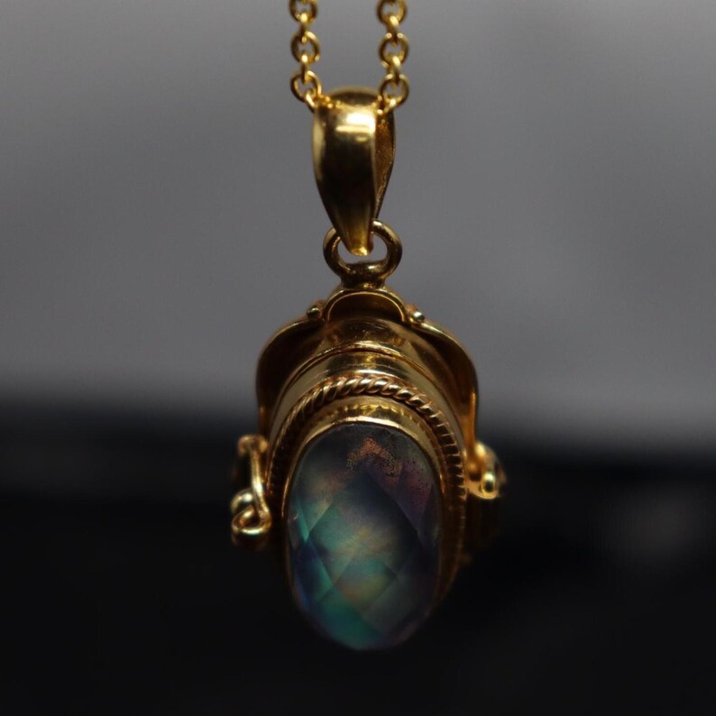 May include: A gold-colored pendant necklace with a large, oval-shaped, iridescent gemstone. The pendant has intricate detailing and a decorative bail. The gemstone displays a range of colors, including blue, green, and purple. The chain is delicate and also gold-colored.