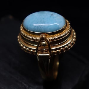 Pode incluir: Um anel de ouro com uma grande pedra preciosa oval azul claro. O anel tem um design detalhado e em camadas com uma faixa texturizada e uma configuração decorativa. O anel está sobre um fundo escuro.