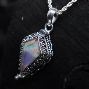 Pu&ograve; includere: Una collana con pendente in argento con una gemma geometrica e iridescente. Il pendente &egrave; incastonato in una montatura in argento dettagliata e strutturata con un bordo a forma di corda. La catena &egrave; una delicata corda d'argento intrecciata.