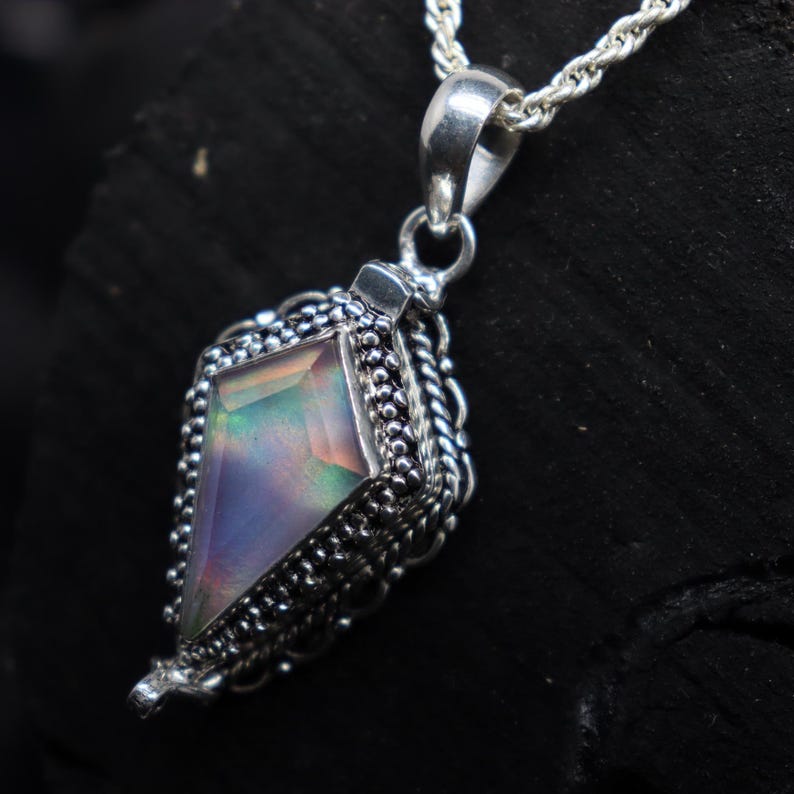 Pu&ograve; includere: Una collana con pendente in argento con una gemma geometrica e iridescente. Il pendente &egrave; incastonato in una montatura in argento dettagliata e strutturata con un bordo a forma di corda. La catena &egrave; una delicata corda d'argento intrecciata.