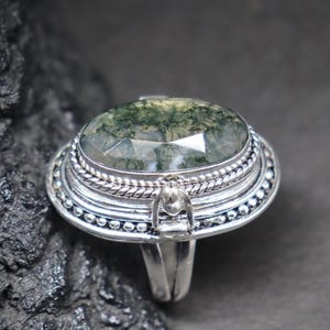 Puede incluir: Anillo de plata con una piedra preciosa ovalada con inclusiones verdes y blancas. El anillo tiene un engaste detallado y ornamentado con un borde trenzado y pequeños detalles de cuentas. La piedra preciosa es el punto focal, sobre un fondo oscuro.