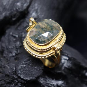 May include: A gold-colored ring with a large, faceted, moss agate stone. The stone is a mix of green and clear colors. The ring has a detailed, ornate design around the stone and on the band. The ring is set against a dark, textured background.