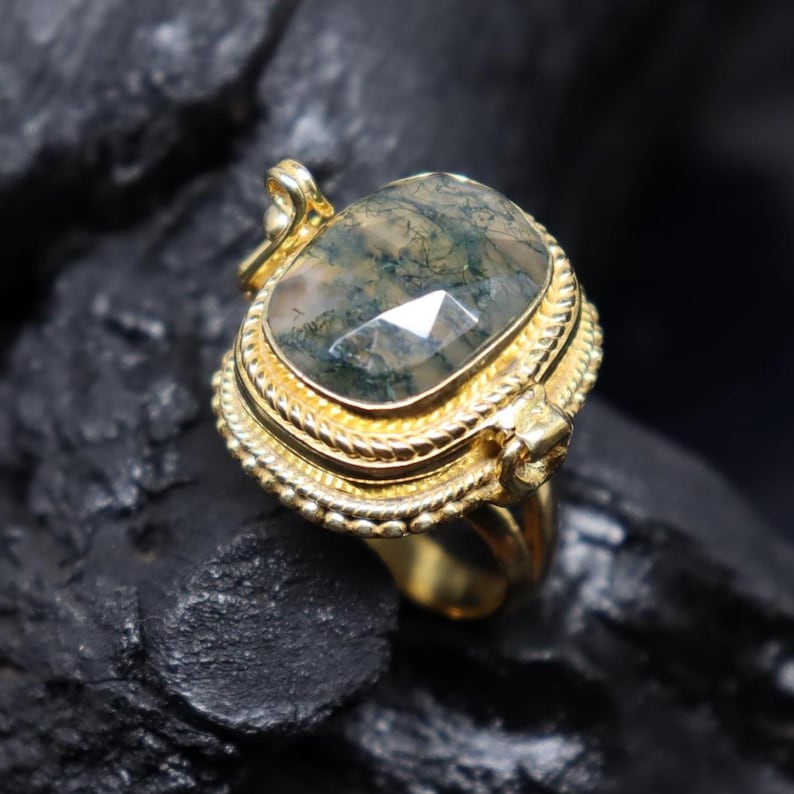 May include: A gold-colored ring with a large, faceted, moss agate stone. The stone is a mix of green and clear colors. The ring has a detailed, ornate design around the stone and on the band. The ring is set against a dark, textured background.