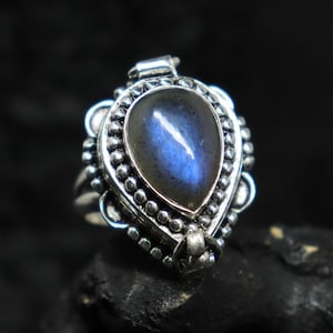 May include: A silver ring with a teardrop-shaped, iridescent blue gemstone. The ring has a detailed, ornate setting with small, round embellishments around the gemstone and on the band. The ring is set against a dark background.