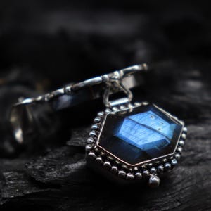 May include: A silver pendant with a faceted, blue gemstone. The gemstone is hexagonal and surrounded by a beaded silver border. The pendant is attached to a silver ring.