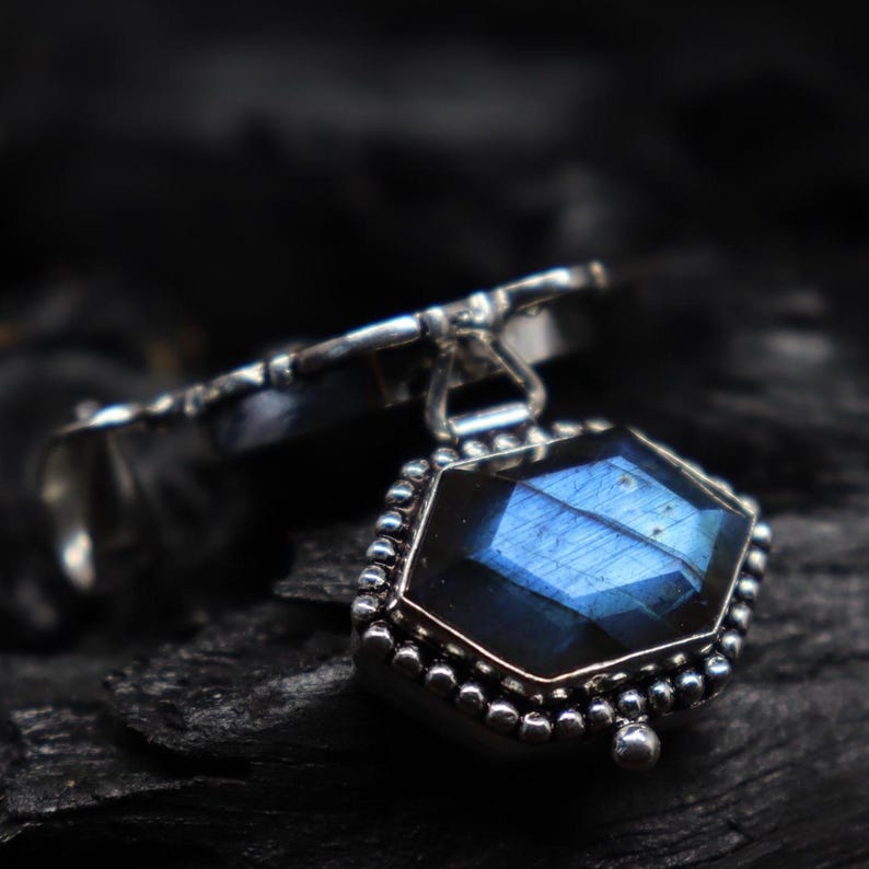 May include: A silver pendant with a faceted, blue gemstone. The gemstone is hexagonal and surrounded by a beaded silver border. The pendant is attached to a silver ring.