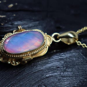 May include: A gold-colored pendant necklace with an oval-shaped, iridescent gemstone. The gemstone displays a blend of pink, blue, and purple hues. The pendant is intricately designed with a decorative border and is suspended from a delicate chain.