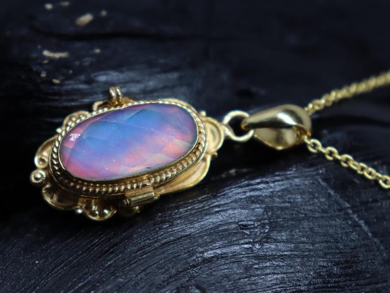 May include: A gold-colored pendant necklace with an oval-shaped, iridescent gemstone. The gemstone displays a blend of pink, blue, and purple hues. The pendant is intricately designed with a decorative border and is suspended from a delicate chain.