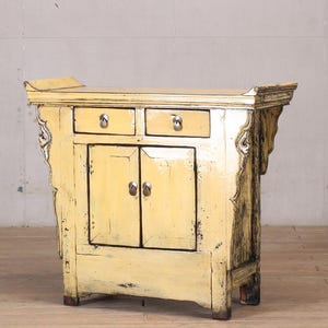 Puede incluir: Un mueble amarillo desgastado con dos cajones pequeños encima de dos puertas de armario. El mueble tiene detalles laterales ornamentados y herrajes plateados. La parte superior tiene un ligero voladizo y la pieza tiene una estética vintage.