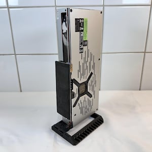 K&ouml;nnte beinhalten: Eine silberne und schwarze Computerkomponente, m&ouml;glicherweise eine Grafikkarte, wird vertikal auf einem schwarzen St&auml;nder angezeigt. Die Komponente hat ein perforiertes Design mit einem zentralen X-f&ouml;rmigen Muster und vertikalen Linien.