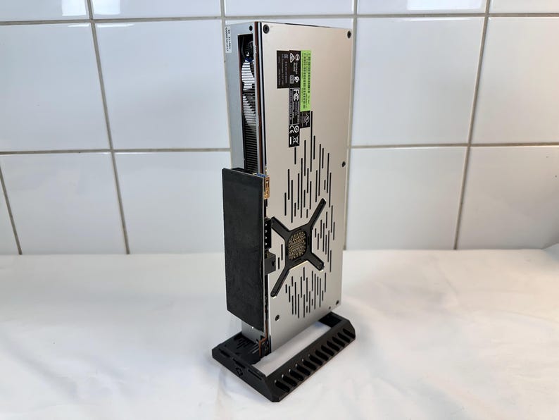 K&ouml;nnte beinhalten: Eine silberne und schwarze Computerkomponente, m&ouml;glicherweise eine Grafikkarte, wird vertikal auf einem schwarzen St&auml;nder angezeigt. Die Komponente hat ein perforiertes Design mit einem zentralen X-f&ouml;rmigen Muster und vertikalen Linien.