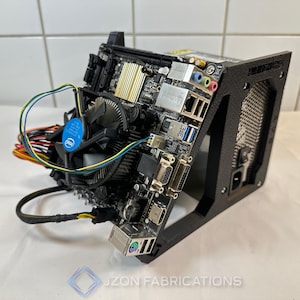 Puede incluir: Una caja de ordenador negra con una placa base, un ventilador de CPU y varios puertos. La placa base tiene disipadores de calor dorados y un ventilador Intel azul. La caja tiene un panel lateral de malla y el texto "JZON FABRICATIONS" en la parte inferior.