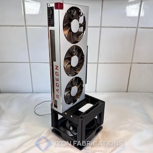 Puede incluir: Una tarjeta gráfica Radeon plateada y roja con tres ventiladores, montada verticalmente sobre un soporte negro. El soporte tiene una base y una sección superior para sujetar la tarjeta. El texto "Radeon" es visible.