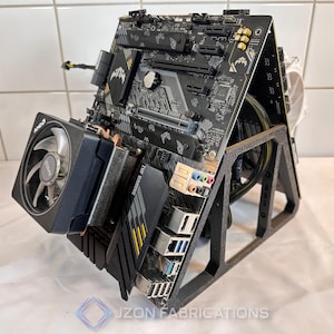 Puede incluir: Una placa base de ordenador negra con un enfriador de CPU y una tarjeta gráfica, montada en un soporte negro. La placa base tiene detalles en amarillo y el texto "TUF GAMING". El soporte tiene el texto "JZON FABRICATIONS".