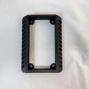 K&ouml;nnte beinhalten: Ein schwarzer rechteckiger Rahmen mit einem strukturierten, geriffelten Design an den Seiten. Der Rahmen hat eine rechteckige &Ouml;ffnung in der Mitte und Befestigungsl&ouml;cher an jeder Ecke. Das Objekt ist wahrscheinlich eine Montagehalterung oder ein Geh&auml;use.